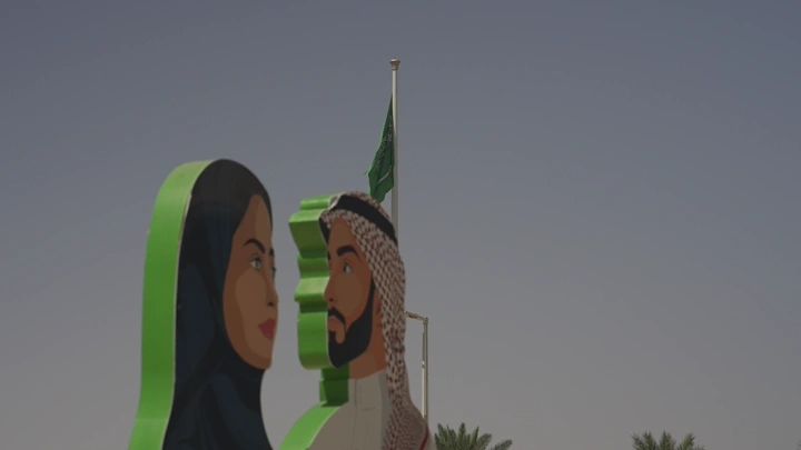 لوحة لشخصين أمام علم سعودي في يوم وطني، لقطة نهارية.