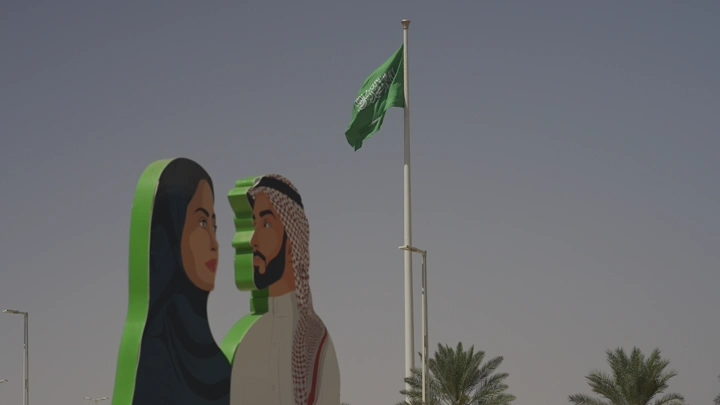علم السعودية يرفرف بجانب لوحة لشخصين، في يوم وطني، لقطة نهارية.