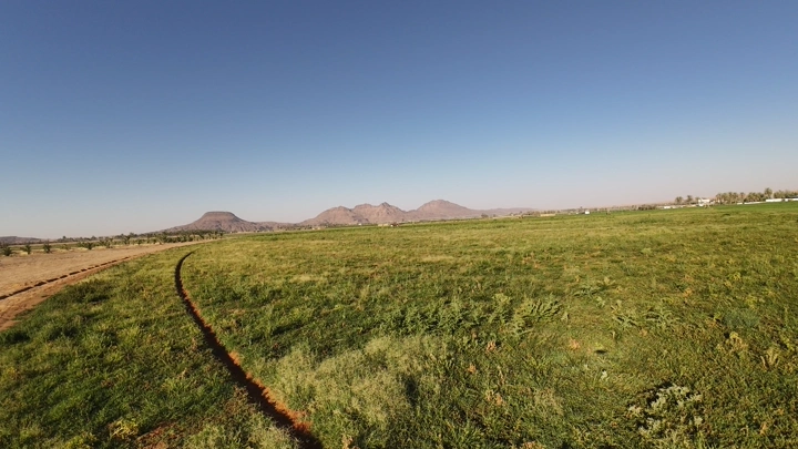 مزرعة خضراء واسعة مع جبال في الخلفية وجرار يحرث الأرض، لقطة بانورامية.
