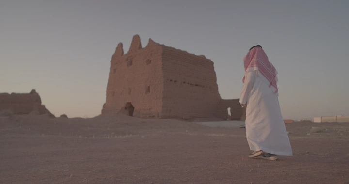 رجل يرتدي الزي السعودي أمام بيت طيني قديم، لقطة نهارية.