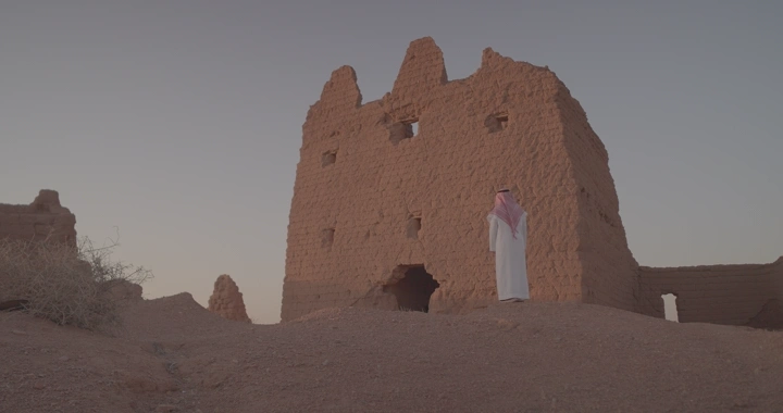 رجل يقف عند بيت طيني قديم في الصحراء عند الغروب، منظر جانبي.