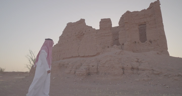 رجل يرتدي الزي السعودي يمشي أمام بيت طيني قديم، لقطة واسعة.
