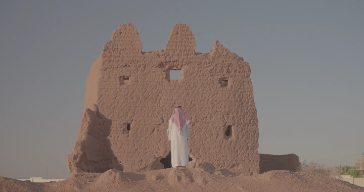 رجل يقف عند بيت طيني في الصحراء تحت سماء زرقاء، لقطة خلفية.