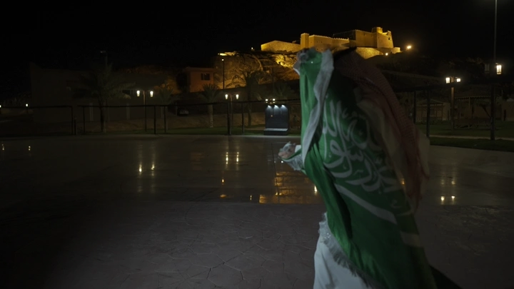 شخص يحمل علم السعودية أمام قلعة عيرف في حائل، لقطة ليلية.