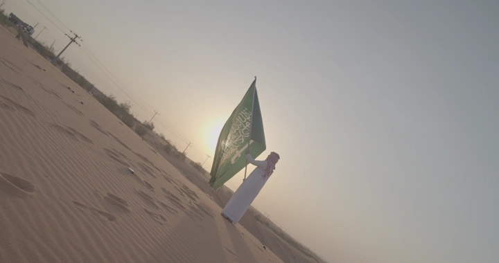 شخص يحمل علم السعودية في الصحراء عند الغروب، لقطة مائلة.