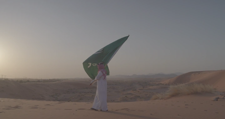 شخص يحمل علم السعودية في النفود عند الغروب، لقطة واسعة.