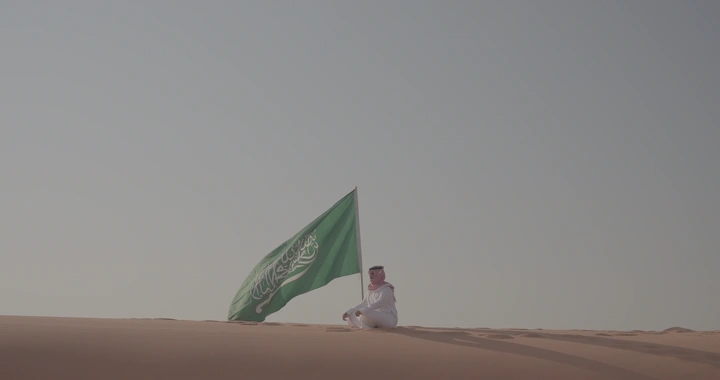 رجل يجلس على كثيب رملي مع علم السعودية، لقطة بعيدة.