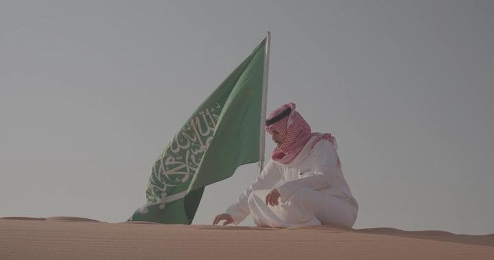 رجل يجلس بجانب علم السعودية في صحراء النفود، لقطة نهارية.