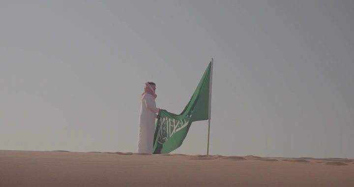 رجل يقف في الصحراء مع علم السعودية، لقطة بعيدة.