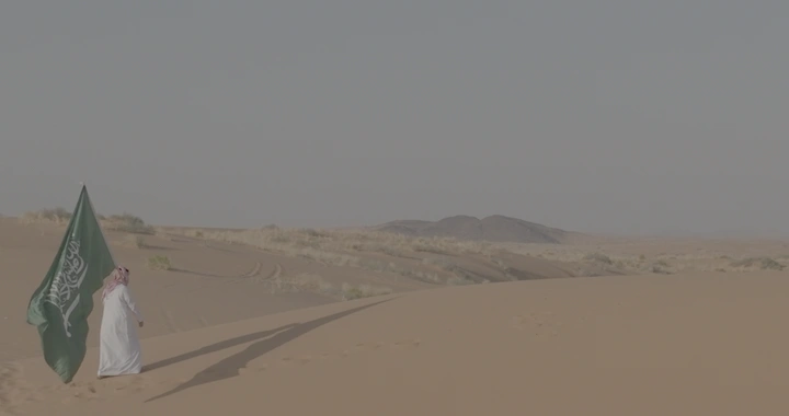 رجل يحمل علم السعودية في صحراء واسعة، لقطة بعيدة.