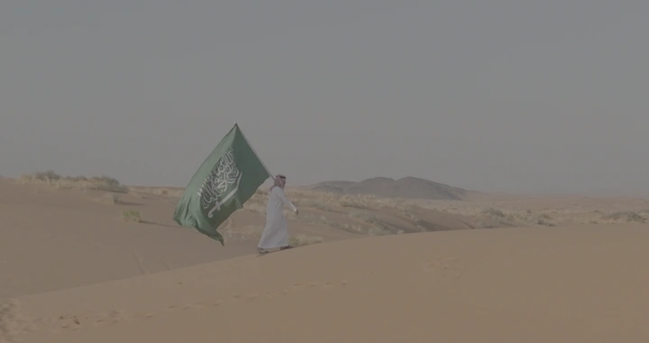 رجل يحمل علم السعودية في صحراء رملية، لقطة بعيدة.