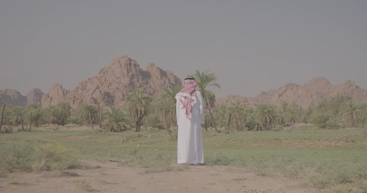 رجل يرتدي ثوبًا في واحة نخيل أمام جبال عقدة، لقطة نهارية.
