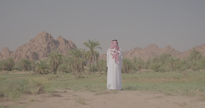 رجل يرتدي ثوبًا تقليديًا يقف أمام جبال عقدة في حائل، لقطة نهارية.