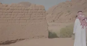 رجل يرتدي الزي التقليدي يقف بجانب بيت طين في الصحراء، لقطة نهارية.