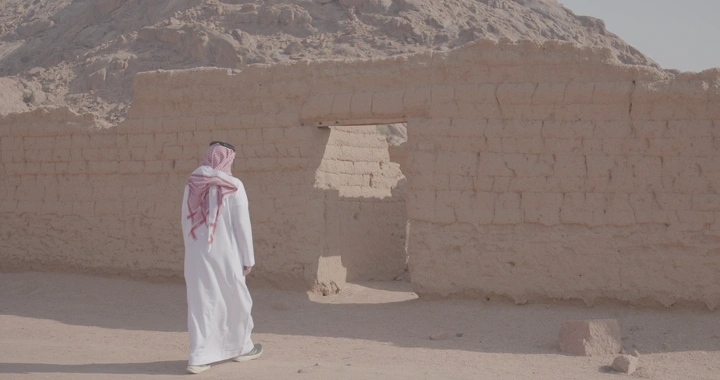 رجل يرتدي ثوبًا أمام بيت طيني في الصحراء، لقطة نهارية.