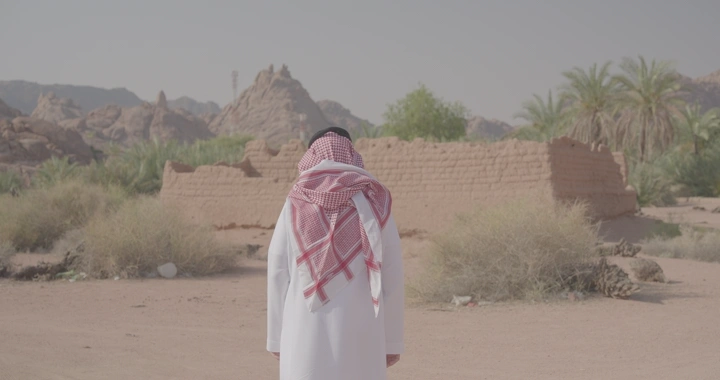 رجل يرتدي ثوبًا وغترة أمام بيت طين في الصحراء، لقطة خلفية.