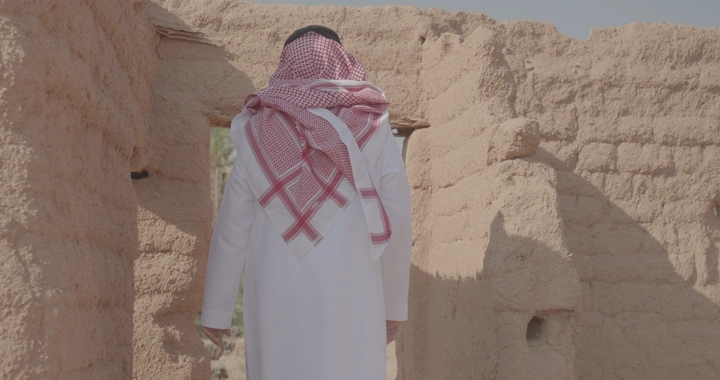 رجل يرتدي ثوب وغترة يقف أمام بيت طيني قديم، لقطة نهارية.