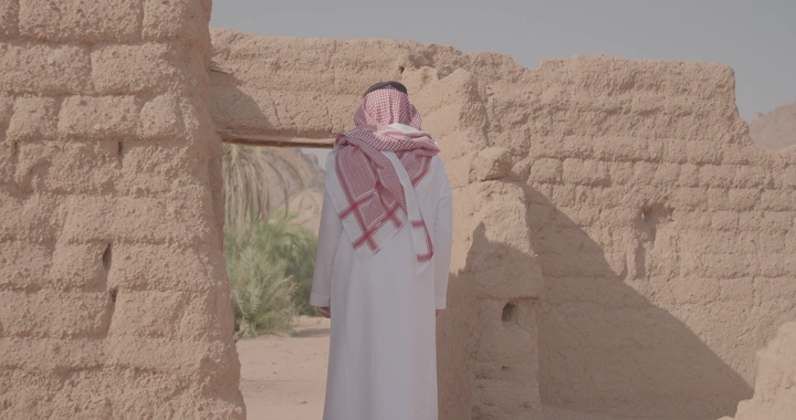 رجل يرتدي الزي التقليدي يقف أمام بيت طين قديم، لقطة نهارية.