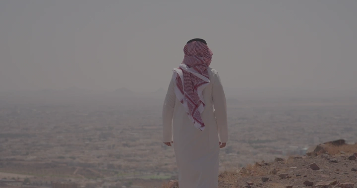 رجل يرتدي الزي التقليدي يقف على تلة تطل على حائل، لقطة خلفية.