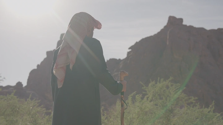 رجل يرتدي غترة يمشي أمام جبال في النهار، لقطة خلفية.