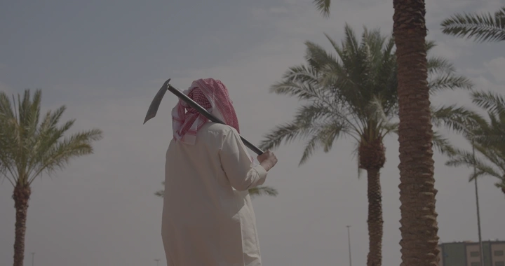 رجل يرتدي ثوبًا ويحمل معولًا في حقل نخيل، لقطة خلفية.