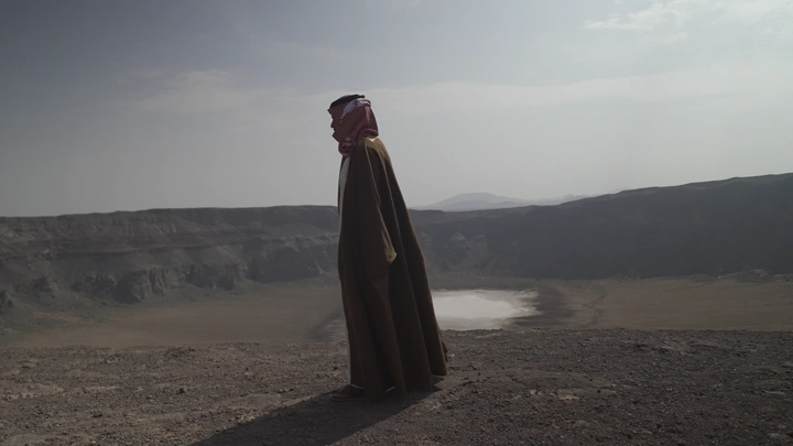 رجل يرتدي الزي السعودي مع البشت يمشي أمام فوهة بركان الهتيمه في حائل، لقطة جانبية.