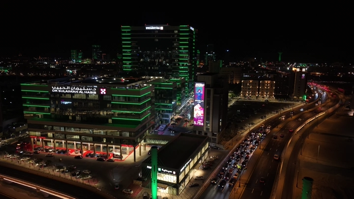 كبري طريق الملك سلمان وطريق الملك فهد بن عبدالعزيز مع إضاءة خضراء على المباني في مدينة الخبر ليلاً، لقطة جوية.