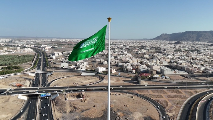 علم السعودية يرفرف فوق مدينة وجبال، تصوير جوي.